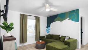 Ein modernes Wohnzimmer mit einer Eckcouch, einem runden Hocker, einem Deckenventilator und einem großen Fenster mit Vorhängen.