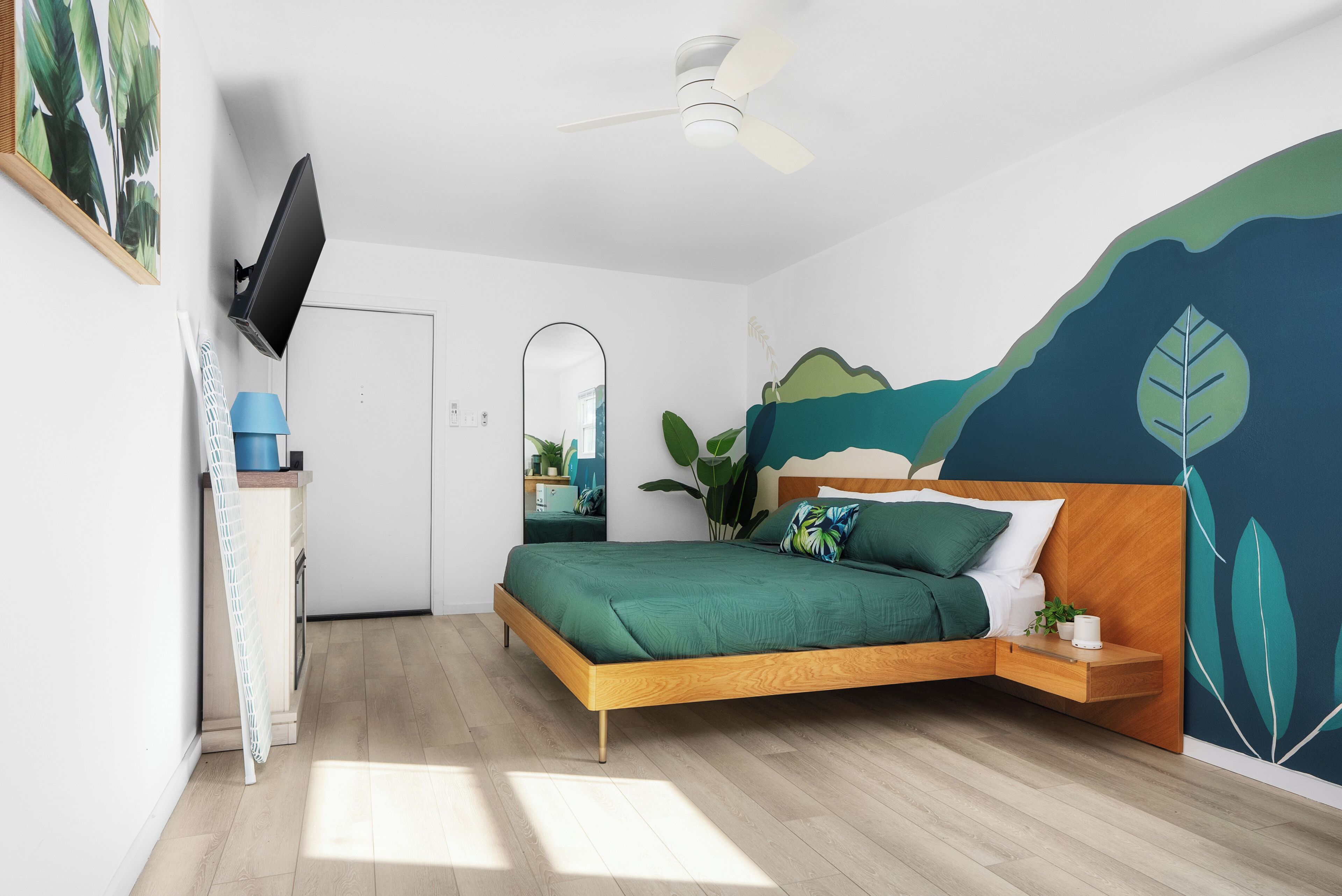 A modern bedroom with a wooden bed, a ceiling fan, a TV mounted on the wall, and a large window allowing natural light.