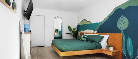 A modern bedroom with a wooden bed, a ceiling fan, a TV mounted on the wall, and a large window allowing natural light.