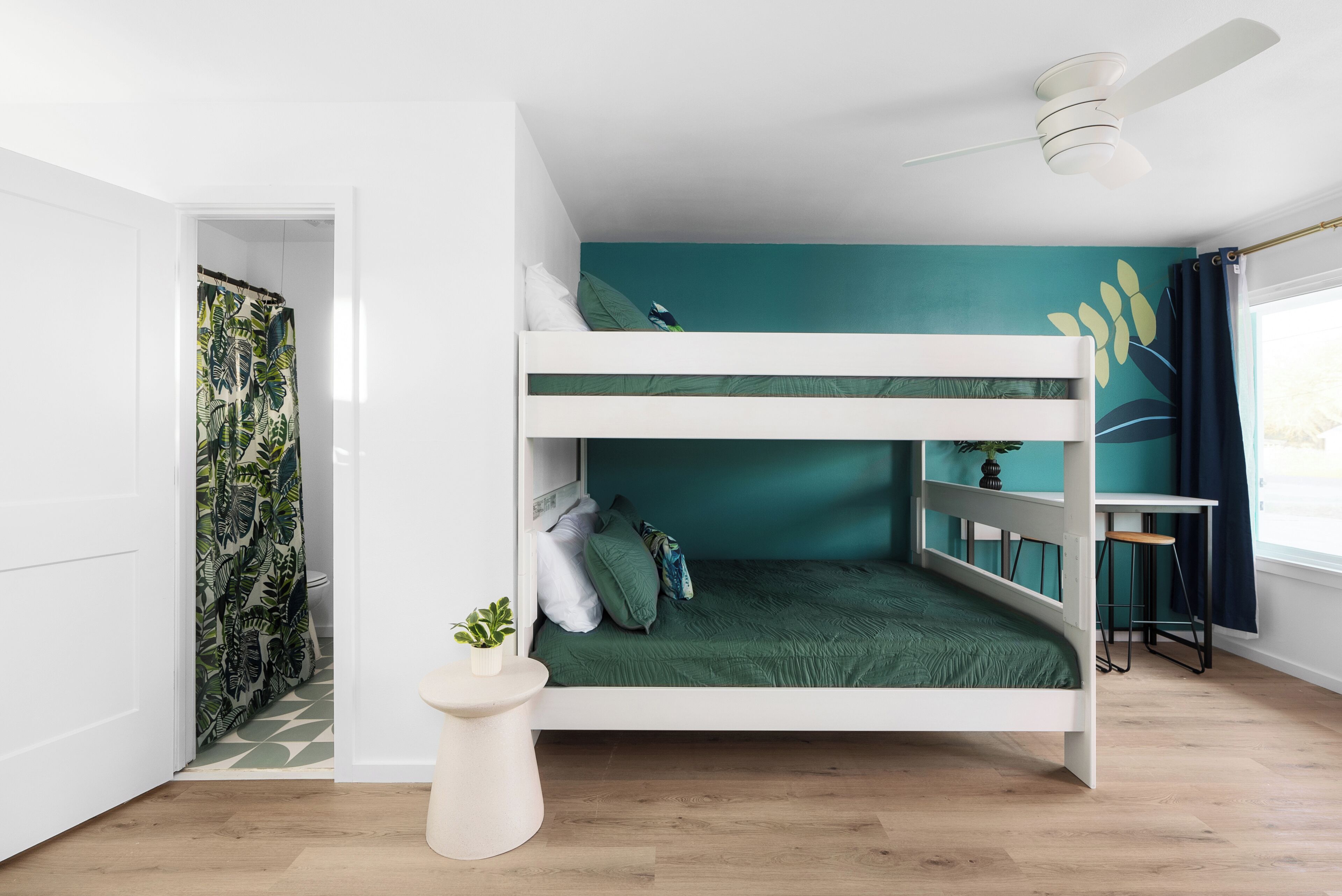 Dormitorio con literas, pared de acento color aguamarina, ventilador de techo y una mesita de noche con una planta.