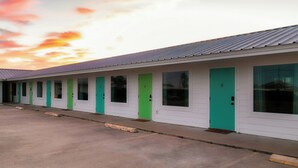 Eine Reihe von Motelzimmern mit grünen Türen und Fenstern, ein Parkplatz und ein Sonnenuntergangshimmel.