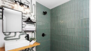 Ein Badezimmer mit weißem Waschbecken, einem Spiegel, einer Dusche mit schwarzem, an der Wand befestigten Duschkopf und einem Wanddesign mit Muster.