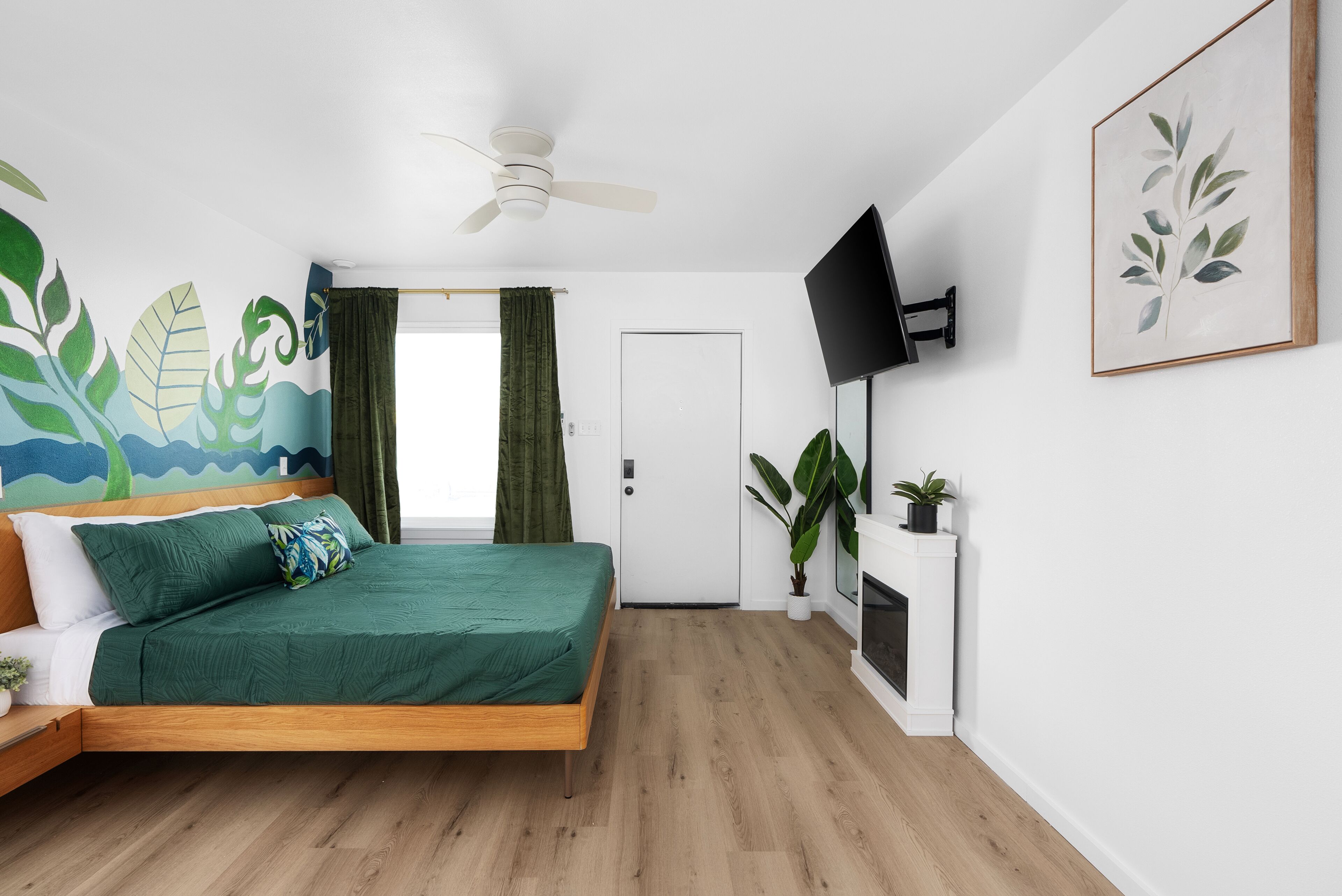 Ein modernes Schlafzimmer mit einem hölzernen Bett, grüner Bettwäsche, einem Deckenventilator, einem Fernseher und einem Wandgemälde.