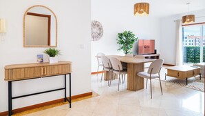 A modern hotel room with a wooden console table, a large mirror, a dining area with a wooden table and chairs, a television, and a large window with a view of buildings.