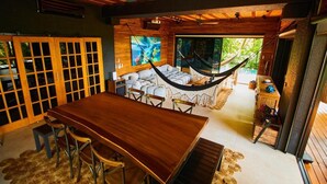 A spacious room with a wooden dining table and chairs, a hammock, and a large window offering a view of the outdoors.