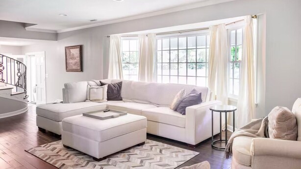 A modern living room with a white sectional sofa, a small ottoman, and a round coffee table. There is a large window with white curtains, a staircase in the background, and a framed picture on the wall.