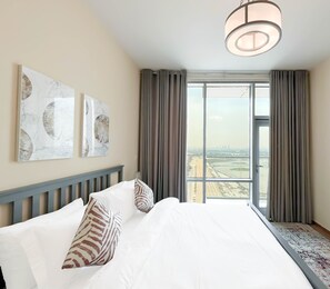 A hotel room with a large bed, two wall-mounted artwork pieces, a modern chandelier, and a view of a cityscape through large windows with curtains. - Sophisticated 2B Apartment at Amna Tower (Dubai)