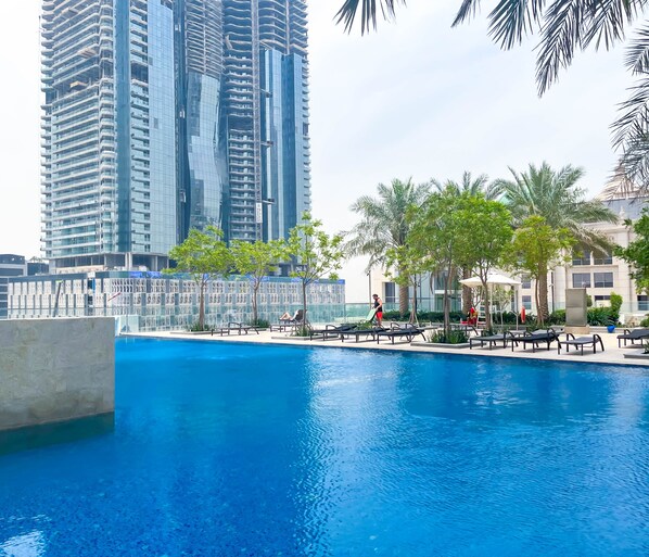 A swimming pool with lounge chairs and palm trees, surrounded by tall buildings. - Sophisticated 2B Apartment at Amna Tower (Dubai)