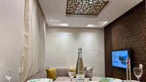 A modern dining area with a table set for two, a bottle of Sidial wine, and a television displaying a scenic view. - Comfort & Style Apartment (Kénitra)