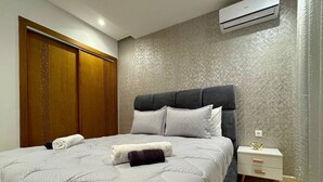 A neatly made bed with a grey quilt, a dark throw pillow, and two rolled towels. There is a white bedside table with a drawer and a small decorative item on top. An air conditioning unit is mounted on the wall above the bed. - Comfort & Style Apartment (Kénitra)