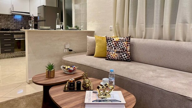 A modern living room with a sofa, coffee table, and a kitchen area in the background. - Comfort & Style Apartment (Kénitra)