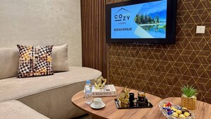 A modern living room with a flat-screen TV displaying a welcome screen, a sofa with a patterned cushion, a coffee table with a book, a tray with snacks, and a decorative pineapple plant. - Comfort & Style Apartment (Kénitra)
