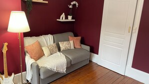 Una habitación con un sofá gris, una puerta blanca, suelo de madera, una lámpara y una guitarra.