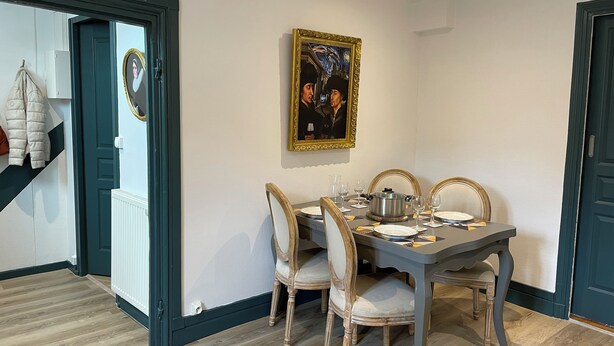 Un comedor con una mesa dispuesta para cuatro, sillas, un cuadro grande enmarco y un perchero con una chaqueta.