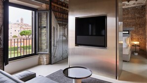 Une chambre moderne dotée d’une grande fenêtre, d’un téléviseur à écran plat fixé sur un mur métallique, d’une table ronde et d’un lit avec une tête de lit.