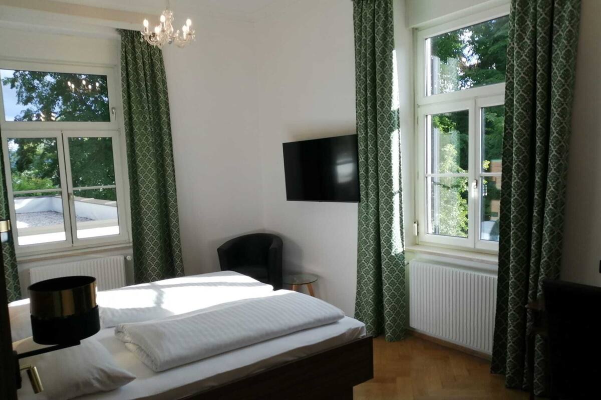 A bedroom with a bed, a chair, a television, and large windows with curtains.