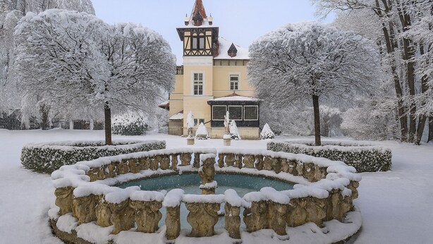 A snow-covered garden with a fountain, a building with a tower, and snow-laden trees. - Glögglhof (Trofaiach)