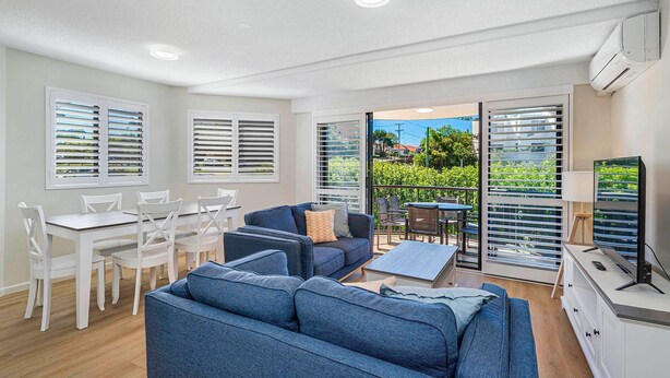 A modern living room with a blue sofa, white chairs, a dining table, and a TV. - Meridian unit 4 Kings Beach QLD (Kings Beach)
