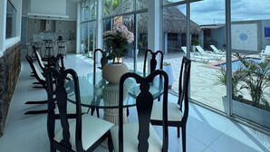 Un amplio comedor interior con una mesa y sillas de vidrio, ventanas grandes que ofrecen vistas a la playa y edificios con techos de paja.