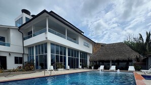 Ein modernes Hotel mit Schwimmbad, einem Reetdachgebäude und weißen Liegestühlen.