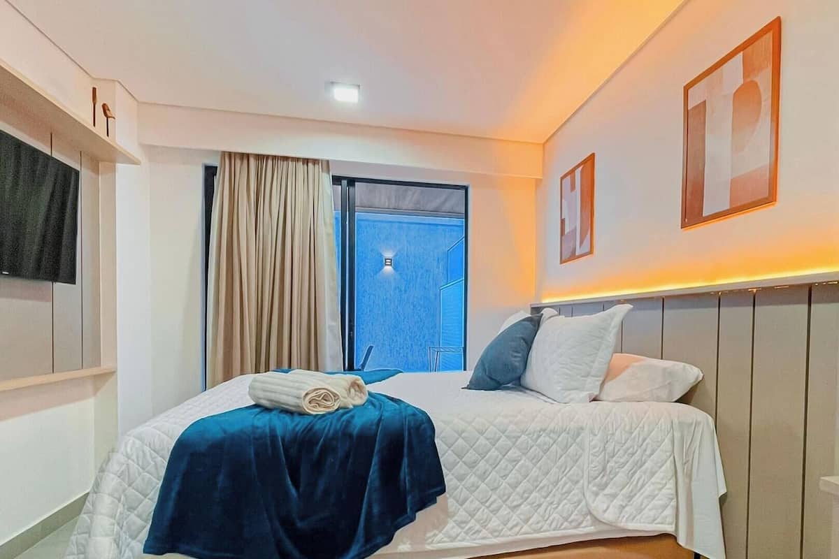 A hotel room with a bed, a TV, a window with curtains, and a wall-mounted shelf.