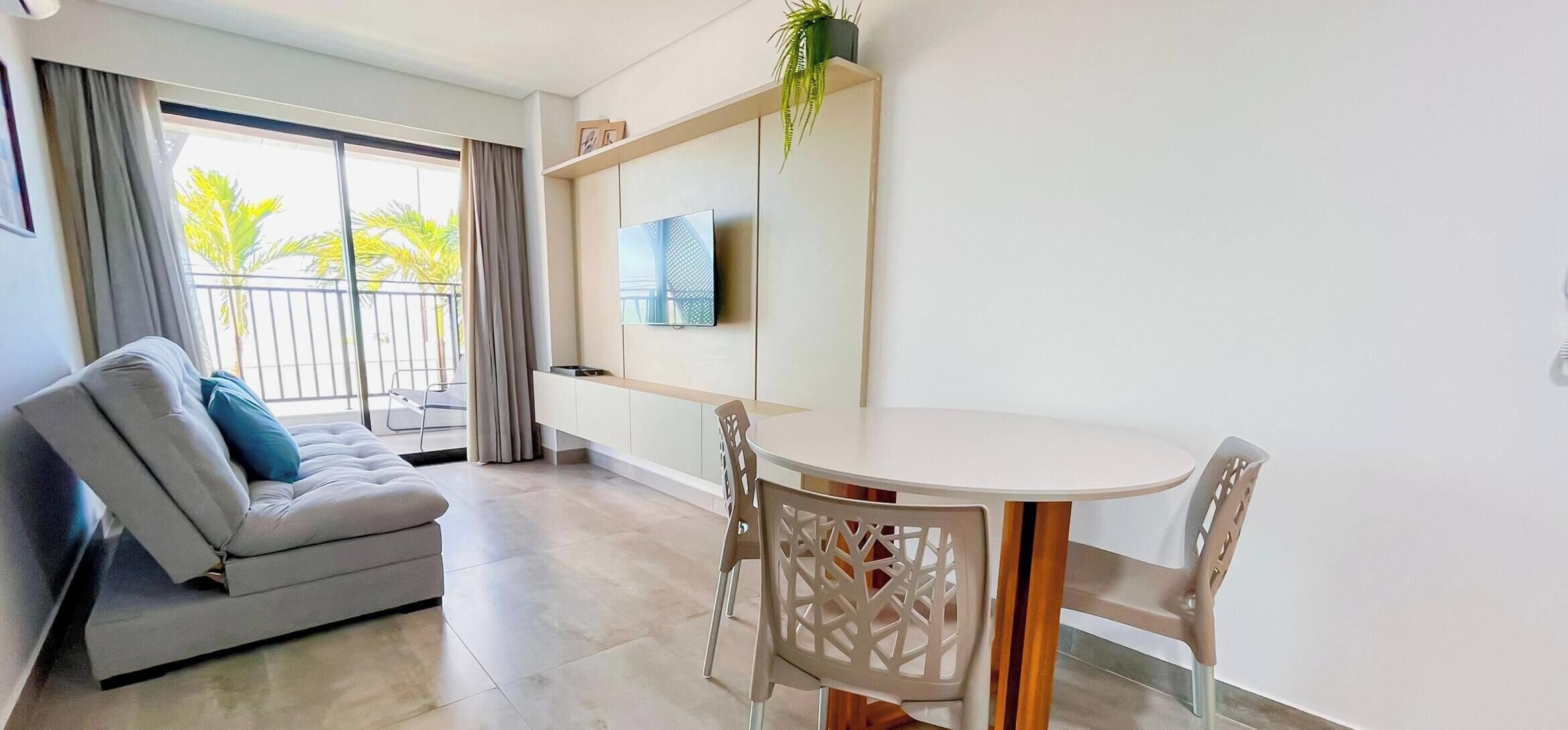 Una sala moderna con un sofá, una mesa de comedor con sillas, un televisor montado en la pared y un balcón con vista a palmeras.