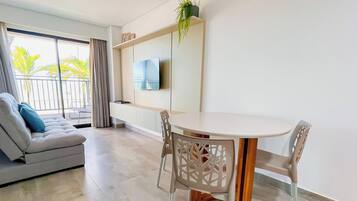 Una sala moderna con un sofá, una mesa de comedor con sillas, un televisor montado en la pared y un balcón con vista a palmeras.