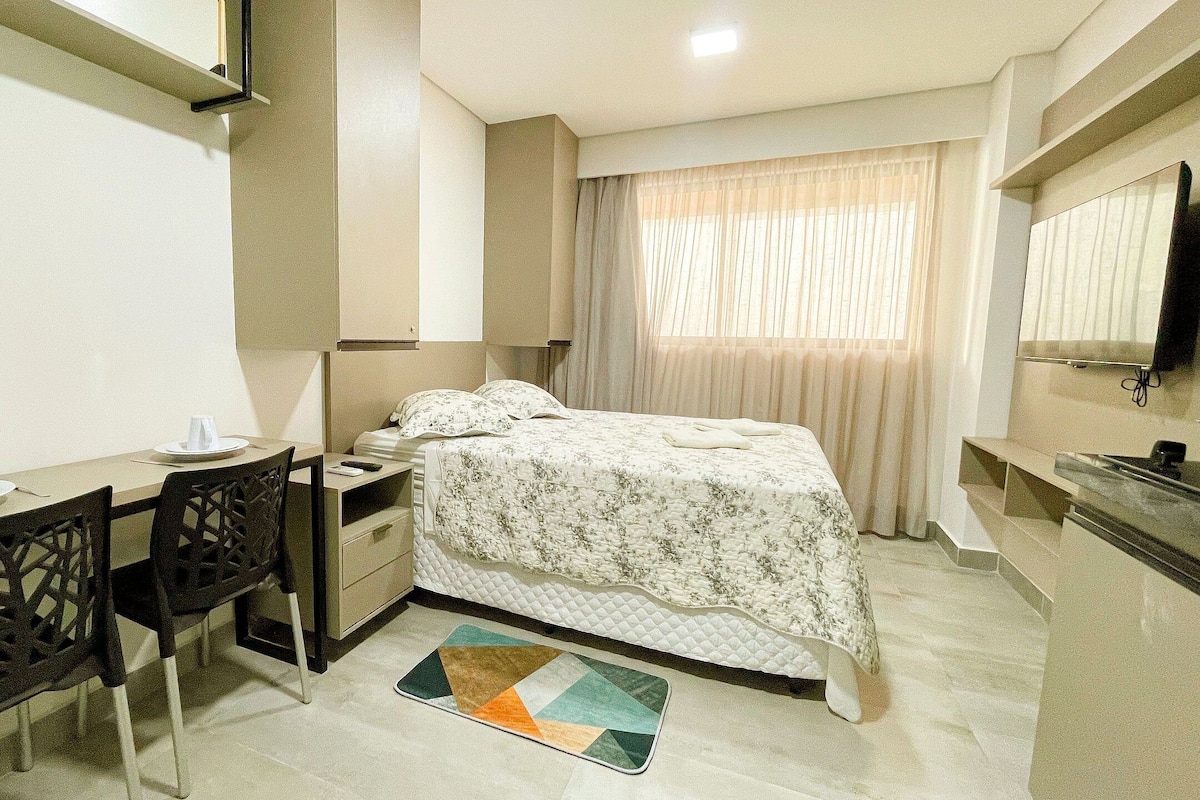 A hotel room with a bed, a desk with chairs, a TV, and a window with curtains.