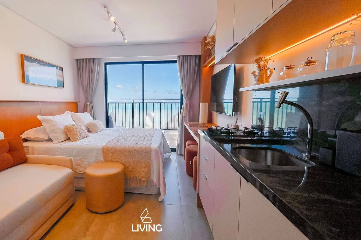 A modern hotel room with a bed, a sofa, a small table, a kitchen area with a stove and sink, and a balcony with a view.