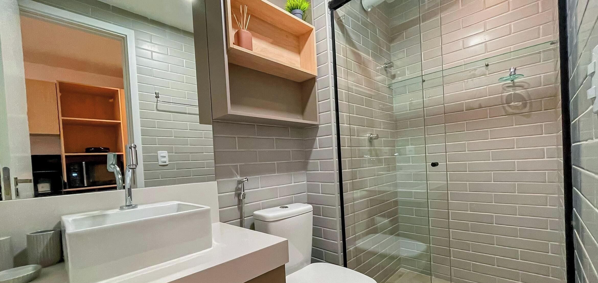 Un baño con un lavamanos blanco, inodoro, cabina de ducha de vidrio y espejo con estante de madera encima.