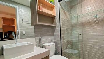 Un baño con un lavamanos blanco, inodoro, cabina de ducha de vidrio y espejo con estante de madera encima.