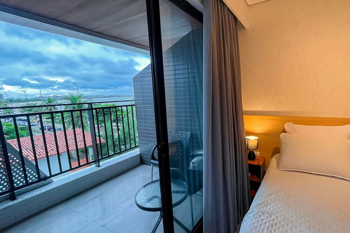 A hotel room with a balcony, a bed with white bedding, a nightstand with a lamp, and a view of the cityscape.