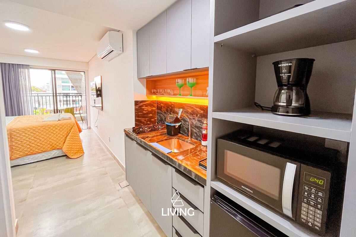 A modern hotel room with a kitchenette, a bed with an orange bedspread, a balcony with a view, and a living area.