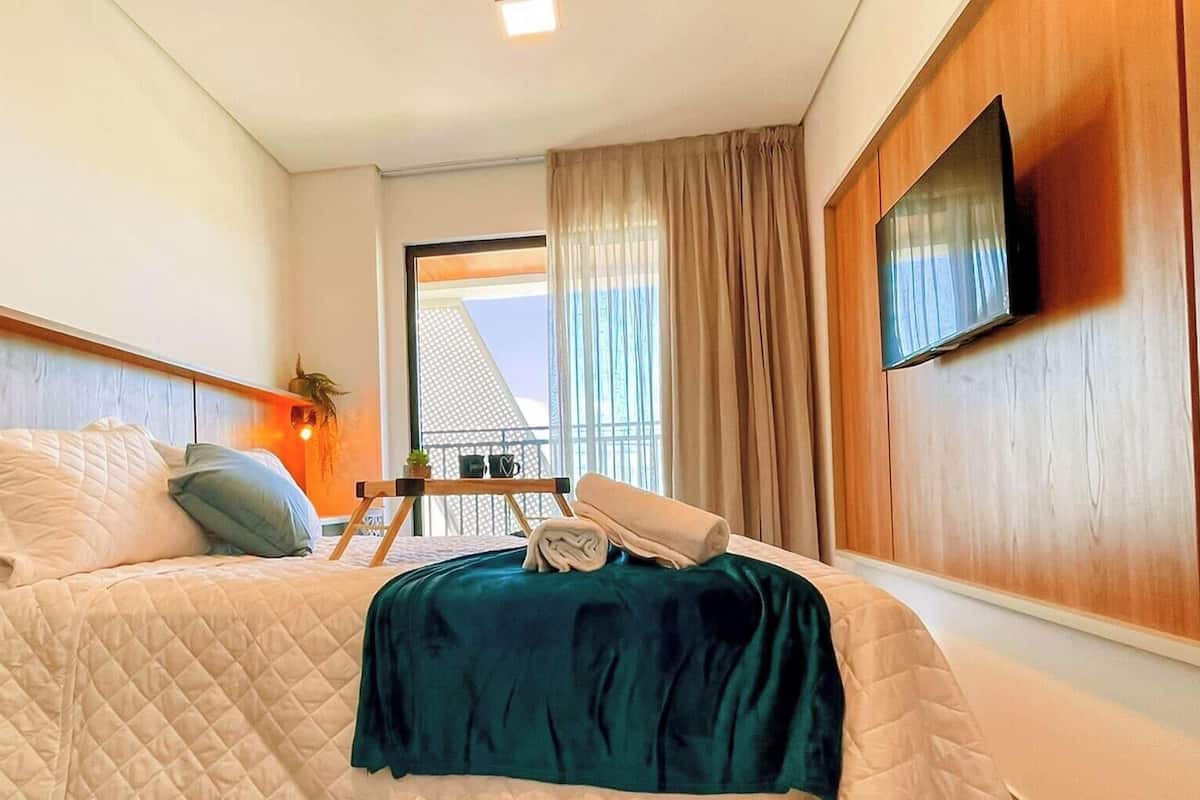A hotel room with a bed, a TV mounted on the wall, a balcony with a view, and a small table with a plant and a cup.