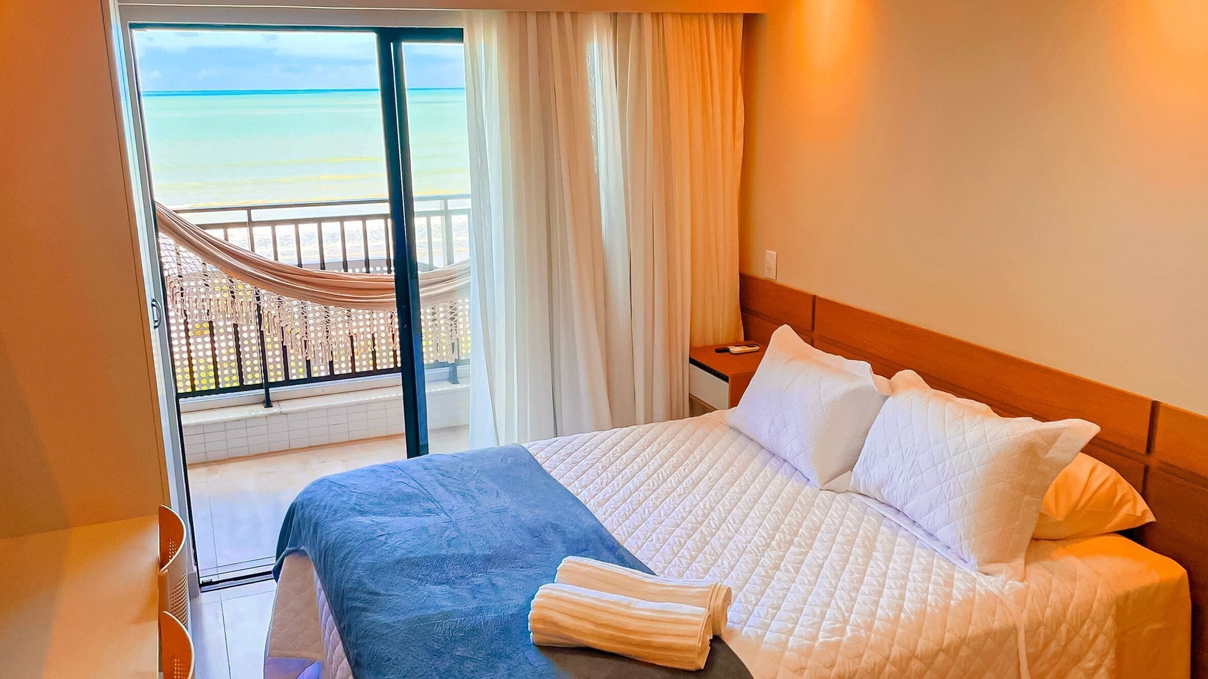 Quarto de hotel com uma cama, mesa de cabeceira, janela com cortinas e vista para a praia.