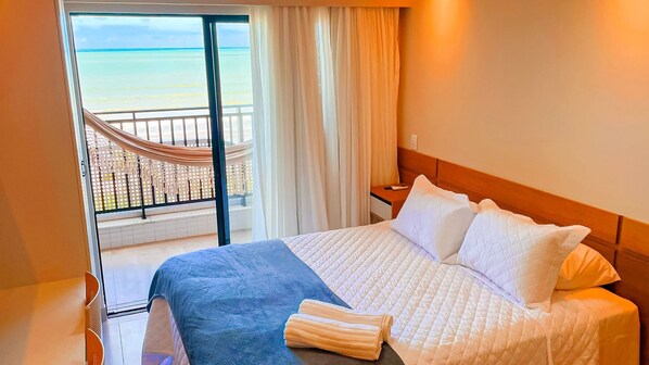 A hotel room with a bed, a nightstand, a window with curtains, and a view of the beach. - Ocean Art Park by Living (Joao Pessoa)