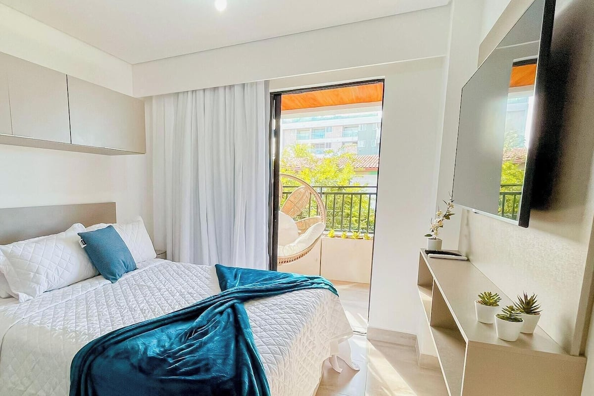 A hotel room with a bed, a TV, a balcony with a view, and a window with curtains.