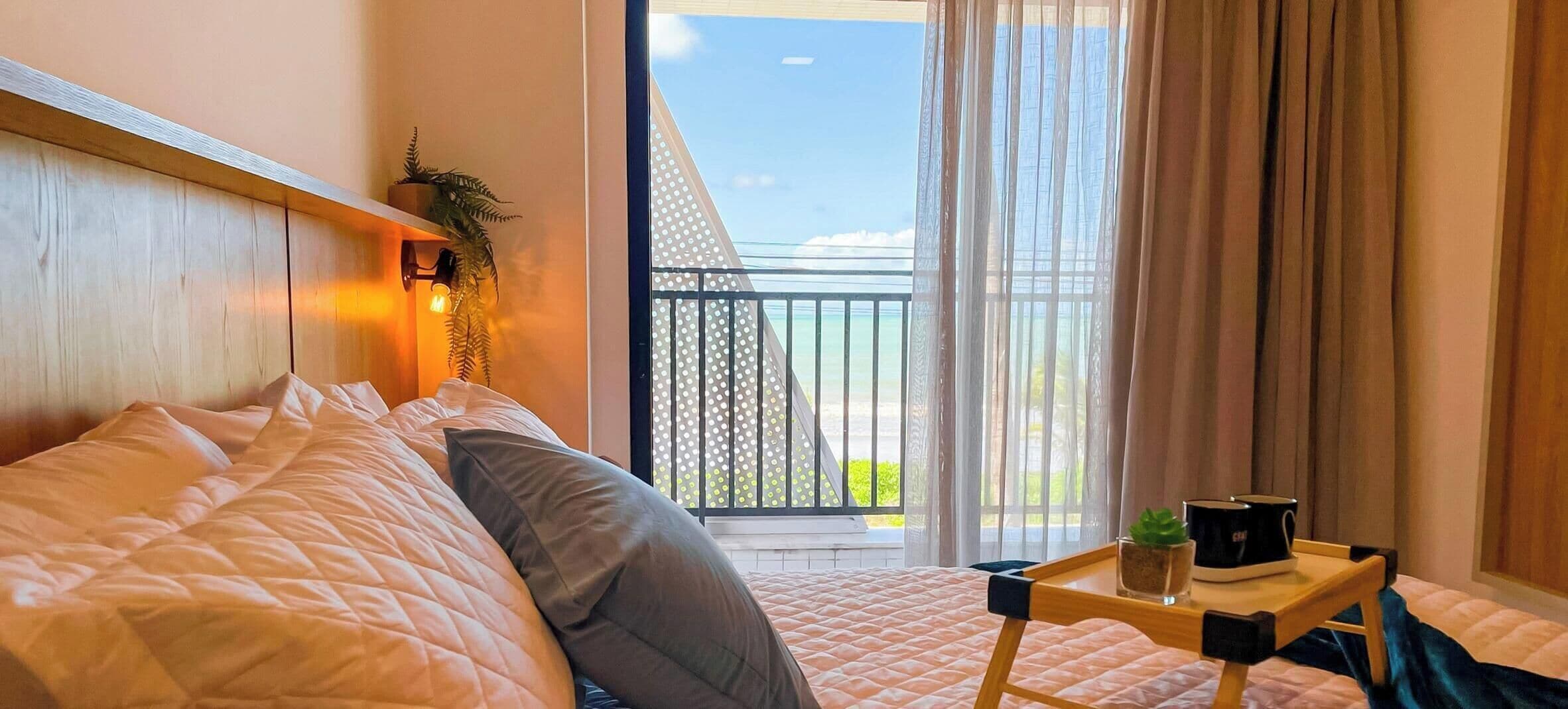 A bedroom with a bed, a bedside table with a plant and a mug, a view of the balcony, and a window with curtains.