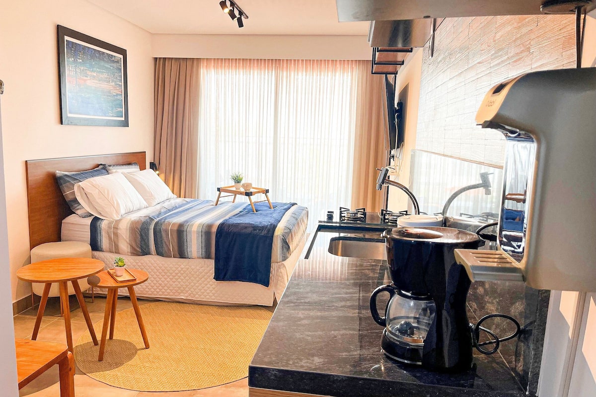 A hotel room with a bed, bedside table, small table, coffee maker, and a view of the city through the window.