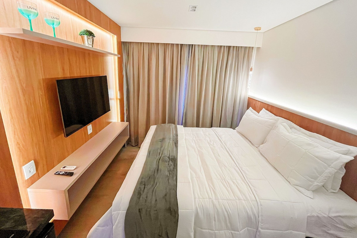 A hotel room with a bed, a TV mounted on the wall, a shelf with decorative items, and a curtain.