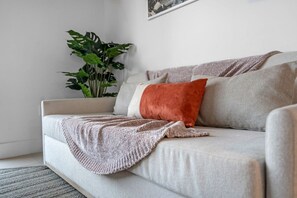 A neutral-toned sofa with a knitted throw blanket and a single orange cushion. A large green plant is visible in the background. - Sunlit 1BR Condo In Downtown Miami (Miami)