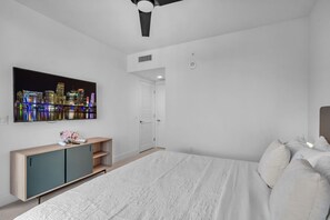 A modern bedroom with a large bed, a TV mounted on the wall, a wooden cabinet, and a door leading to another room. - Bright & Breezy 1 Bed Unit With Water & City Views (Miami)