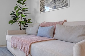 A beige sofa with a pink knitted throw blanket and two pillows, one blue and one beige, against a white wall. A large green plant is visible to the left of the sofa. - Bright & Breezy 1 Bed Unit With Water & City Views (Miami)