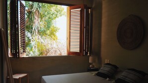A bedroom with a bed, a chair, a nightstand with a lamp, and a window with wooden shutters. - Casa Boga - Comfortable designer house in La Punta (Brisas de Zicatela)