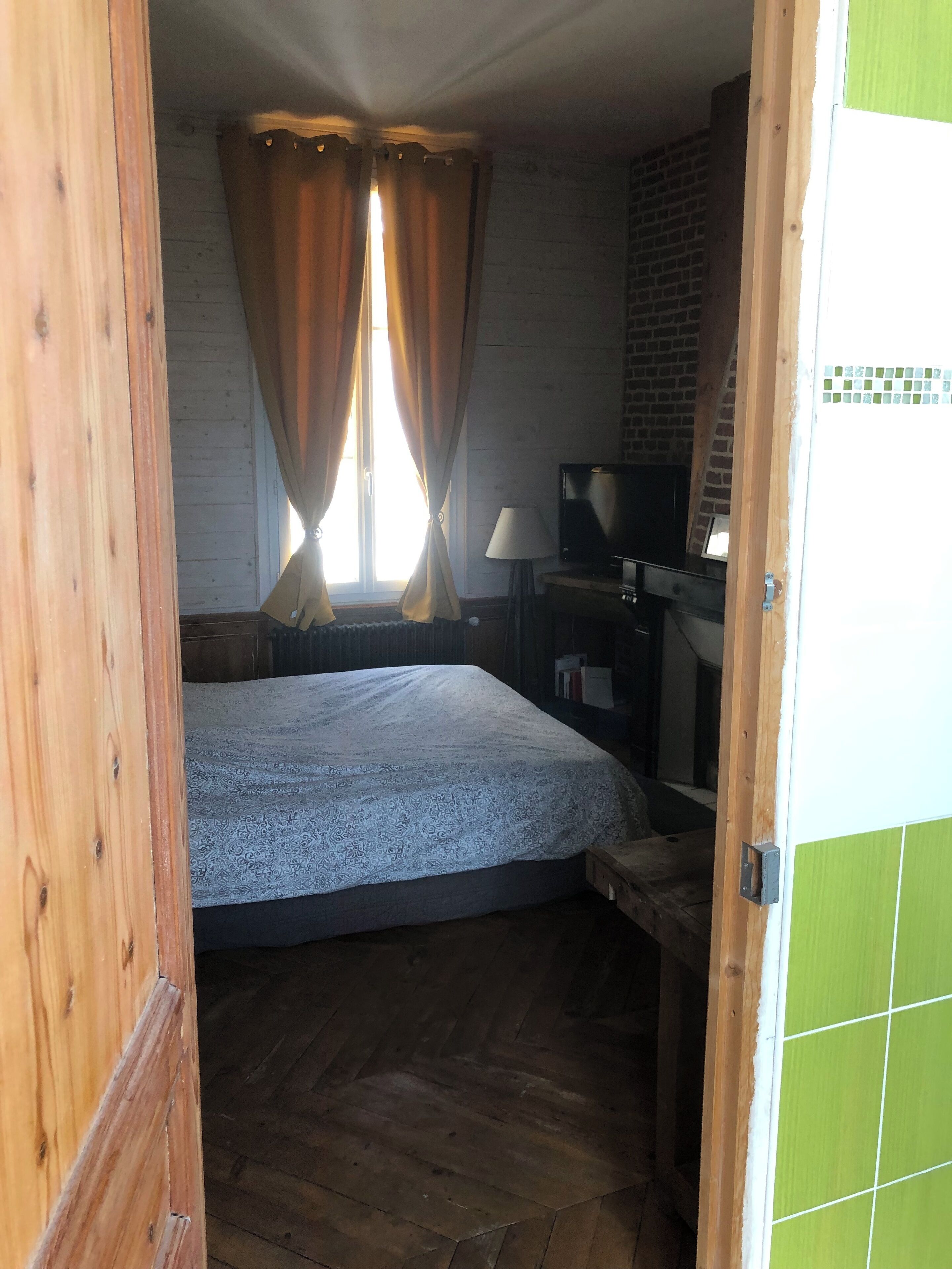 Ein Schlafzimmer mit einem Bett, Holzboden, Vorhängen, einem Schreibtisch mit Lampe und einem Fernseher.