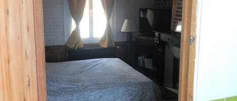 Ein Schlafzimmer mit einem Bett, Holzboden, Vorhängen, einem Schreibtisch mit Lampe und einem Fernseher.