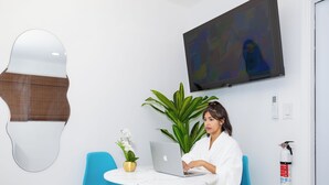 Une pièce avec un mur blanc, un grand miroir, une chaise bleue, une table avec un ordinateur portable, une plante en pot et un extincteur.