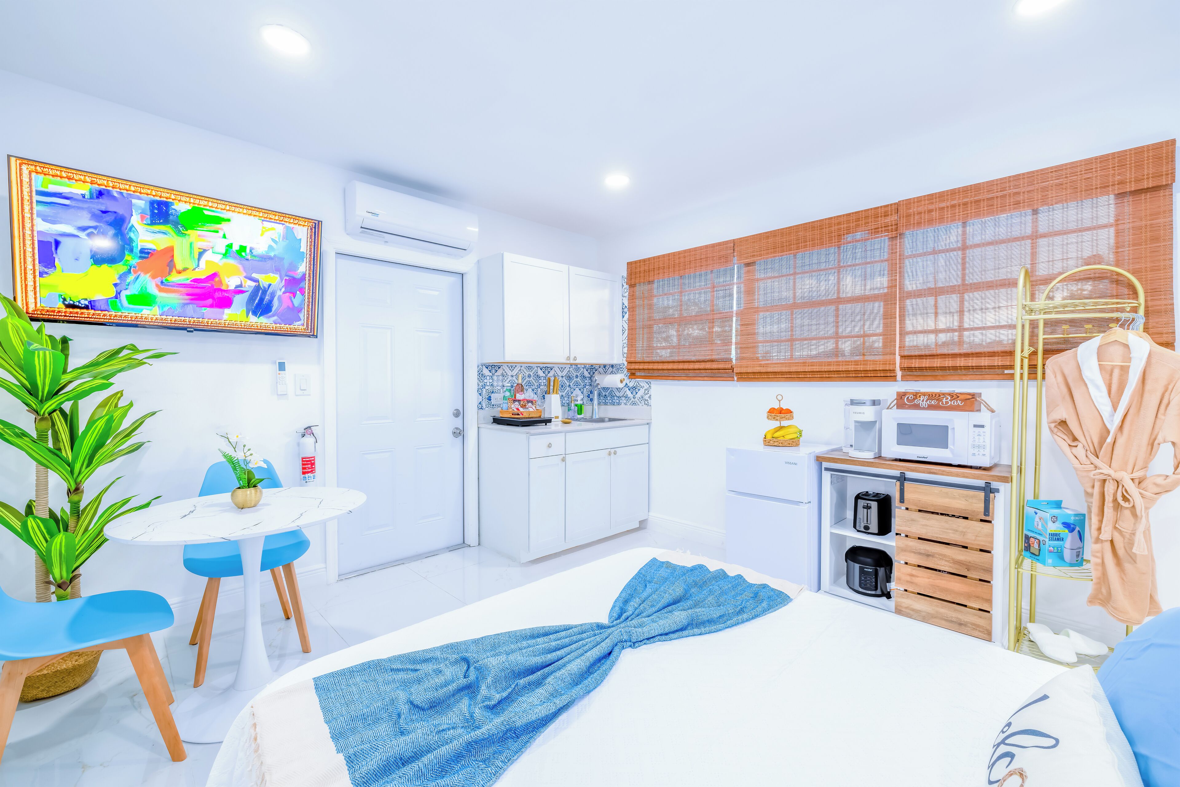 A modern hotel room with a bed, a small dining table, a kitchenette, and a large window with blinds.