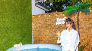 Une personne vêtue d’une longue robe blanche tient un verre de jus d’orange. Elle se tient à côté d’un jacuzzi gonflable, avec un décor en bambou et une pancarte sur laquelle on peut lire : « Soyez l’endroit idéal. »