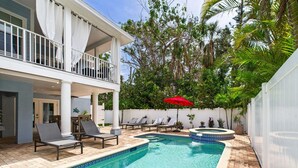 A swimming pool with lounge chairs and a hot tub, surrounded by a white fence and lush greenery.